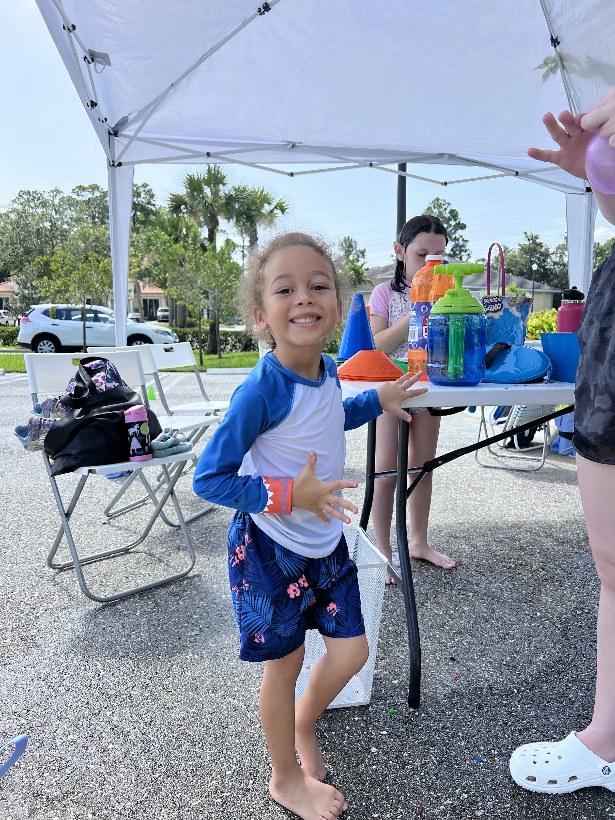 A young girl with curly hair wearing a blue and white long-sleeve shirt and navy shorts with pink flowers, smiling and posing at an outdoor birthday party with water toys and supplies on a table under a white canopy.