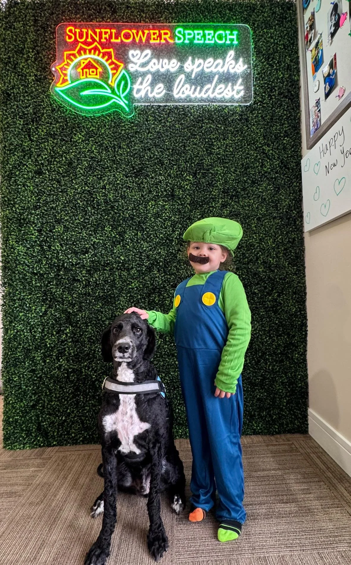Child dressed as Luigi from Super Mario, wearing a green hat and mustache, standing next to a black and white dog in front of a green wall with a bright neon sign that reads 'Sunflower Speech' and 'Love speaks the loudest'.