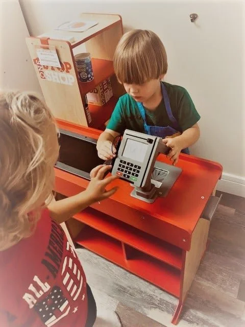 A young boy wearing a green shirt and apron is playing with a toy cash register at a pretend store. A girl is standing in front of him, reaching out to touch the register. The scene is set in a play area with a small wooden shelf behind them.