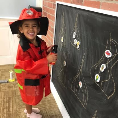 A young girl dressed as a firefighter drawing on a chalkboard with chalk.