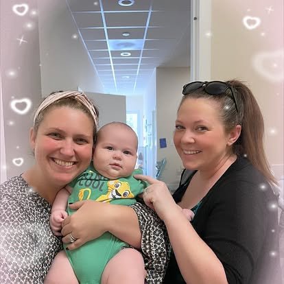 Two women smiling and holding a baby in a hospital corridor, with decorative light effects