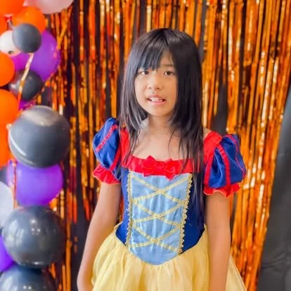 Young girl dressed in a Snow White costume standing in front of Halloween decorations with purple, black, and orange balloons and a shiny orange curtain.