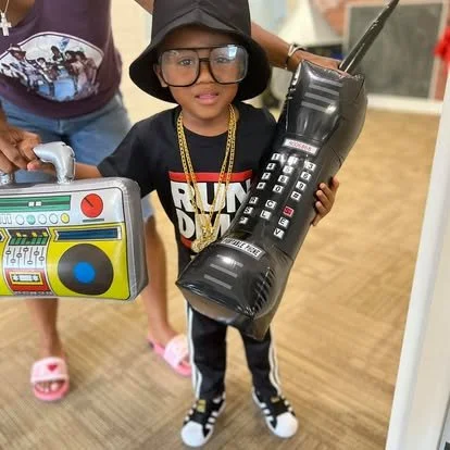 Child wearing glasses and a hat, holding a large black toy telephone, with a smaller colorful boombox next to an adult's hand in the background.