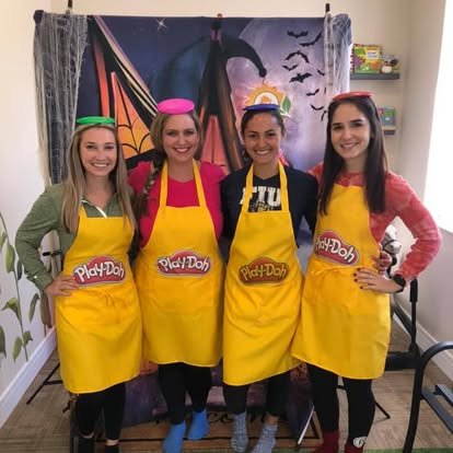 Four women dressed in colorful aprons and headbands posing in front of a Halloween-themed backdrop.