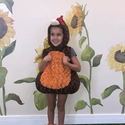 A young girl dressed in a sunflower costume holding an orange plush toy, standing in front of a sunflower mural.