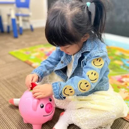 Little girl in a denim jacket with smiley face patches, playing with a pink pig-shaped coin bank.