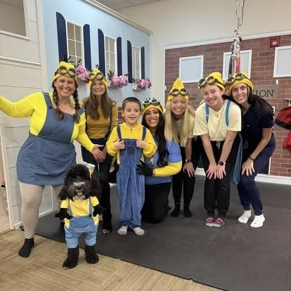 Group of six people dressed as Minions from 'Despicable Me,' with a small dog in a matching Minion costume, in a room with blue and yellow décor and a brick wall.