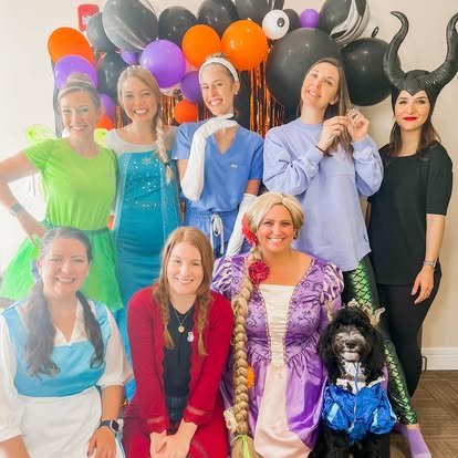 Group of women in colorful costumes and a woman dressed as Rapunzel, with a black and white dog, in front of Halloween or party decorations including black, orange, purple, and pink balloons.