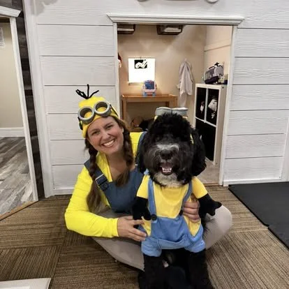 A woman dressed as a minion from Despicable Me, wearing goggles and a yellow shirt, sitting on the floor with a large black dog dressed in a similar minion costume, inside a house with white wooden walls and a doorway.