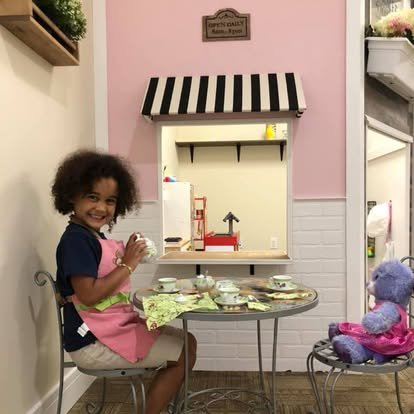 Child with curly hair in an apron sitting at a small table with tea set, a purple stuffed animal on a chair, in a miniature play cafe with pink and white decor.