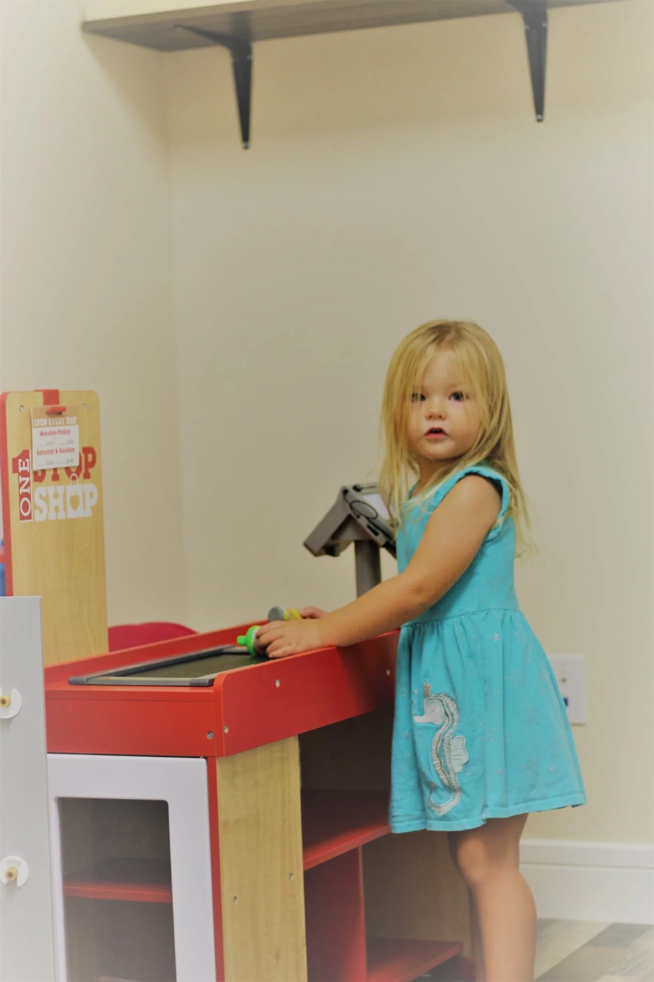 A young girl with blonde hair wearing a blue dress is standing at a red and wood-colored play kitchen, holding a toy spoon, in a room with plain light-colored walls.