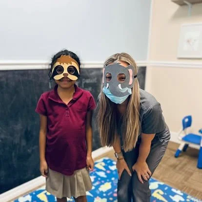 Two children wearing animal face masks, one with a dog mask and the other with a panda mask, in a classroom setting.