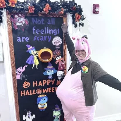 Person in a pig costume standing next to a Halloween-themed chalkboard with characters and decorations.