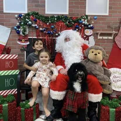 Santa Claus sitting with two children and a dog, surrounded by Christmas decorations and gifts.