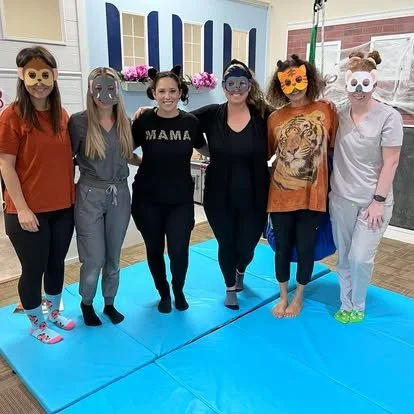 Six women standing on a blue mat, wearing animal-themed masks, with one woman dressed as a tiger, in a room with a pink brick wall and decorative flowers in the background.