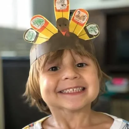 Child wearing a Thanksgiving-themed paper turkey hat with colorful paper feathers.