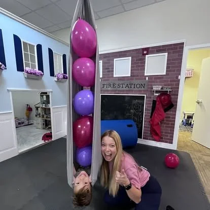 Child hanging upside down with a woman giving a thumbs-up in a playroom with balloons and a fire station themed wall.
