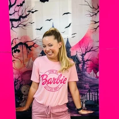 A woman with a ponytail wearing a pink Barbie t-shirt stands in front of a spooky, Halloween-themed backdrop with bats, a full moon, and a haunted house.
