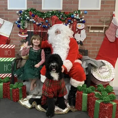 Child in green dress sitting next to Santa Claus and a black dog wearing a red plaid outfit, with Christmas decorations and gifts in the background.