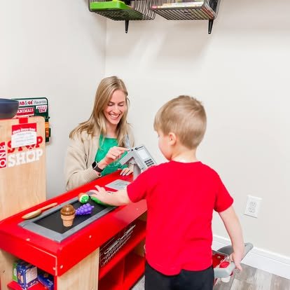 A young woman at a toy checkout stand with a little boy in a red shirt. The woman is smiling as she uses a toy cash register to ring up a toy cupcake. The setting appears to be a play area or store.