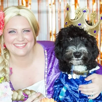 A woman dressed as a queen with a crown, sitting beside a dog also wearing a crown and royal blue cape.
