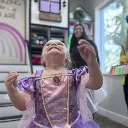 A young girl in a princess dress laughing with her head tilted back, in a room with shelves and colorful toys.