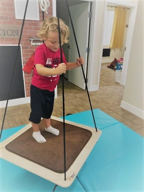 A young boy standing on a swing pad inside a house, holding onto the swing ropes and smiling.