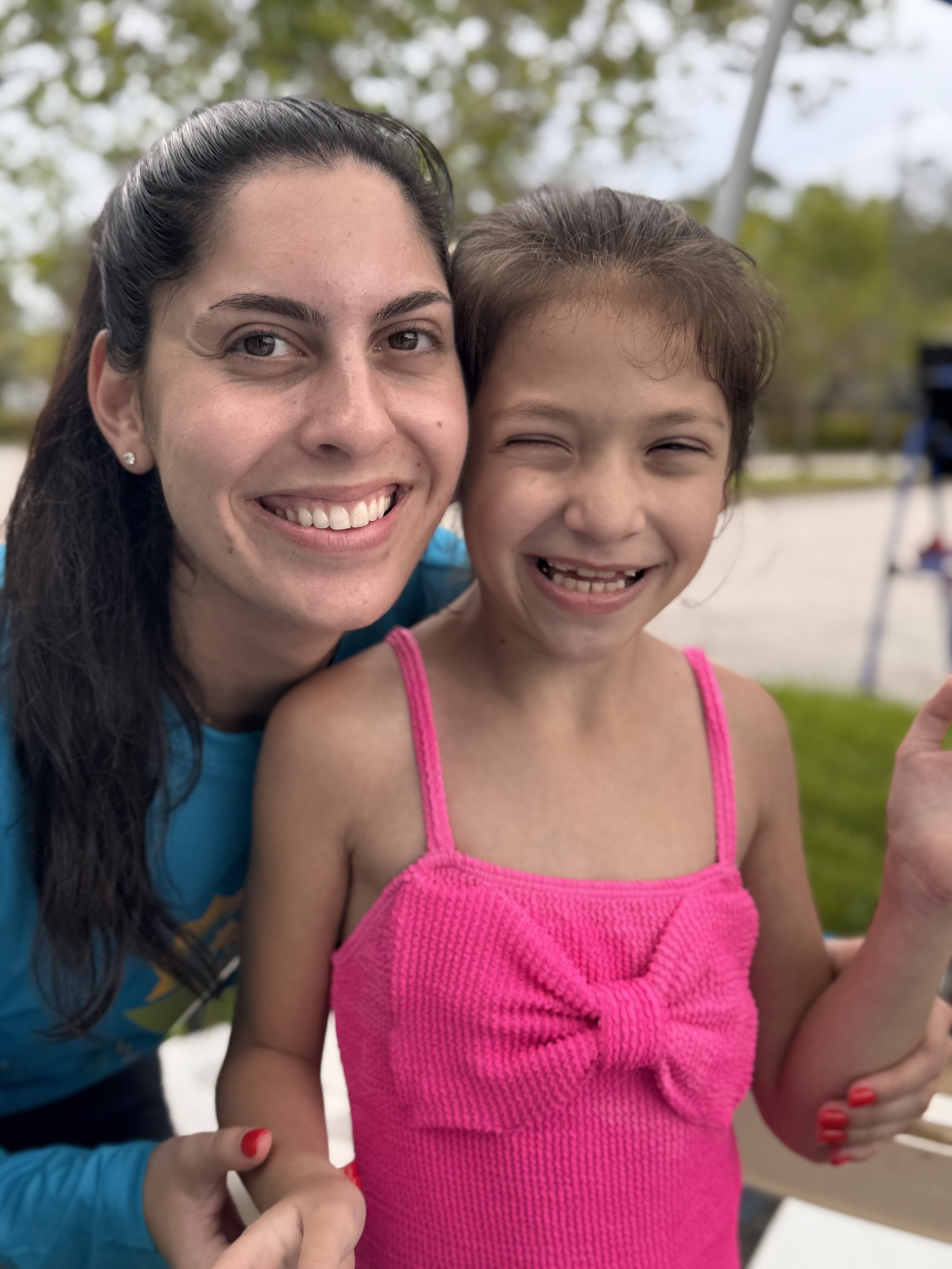 A woman and a young girl smiling outdoors, close together, with a playground in the background.