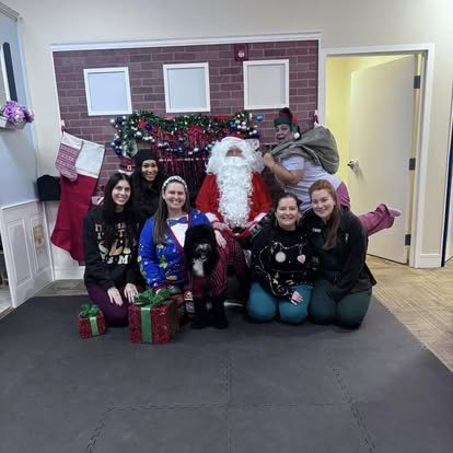 Group of six young women in winter clothing, sitting and kneeling on a black carpeted floor, posing with Santa Claus and a black and white dog in front of a festive Christmas display. The display includes a large red stocking, a decorated garland with ornaments and lights, and a faux brick wall backdrop. A woman on the right is holding a child dressed in a brown coat and hat. Several wrapped gift boxes are on the floor in front.