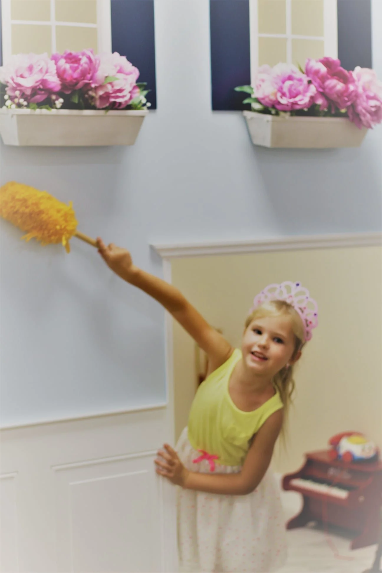 A young girl with a pink crown dolling a yellow top and a pink and white skirt, leaning out of a doorway, holding a duster in the air, with pink flowers in window boxes above her.