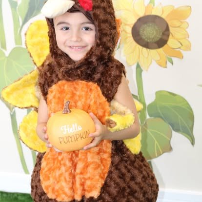 Child in a bee costume holding a small pumpkin with 'Hello Pumpkin' written on it, standing in front of sunflower and greenery backdrop.