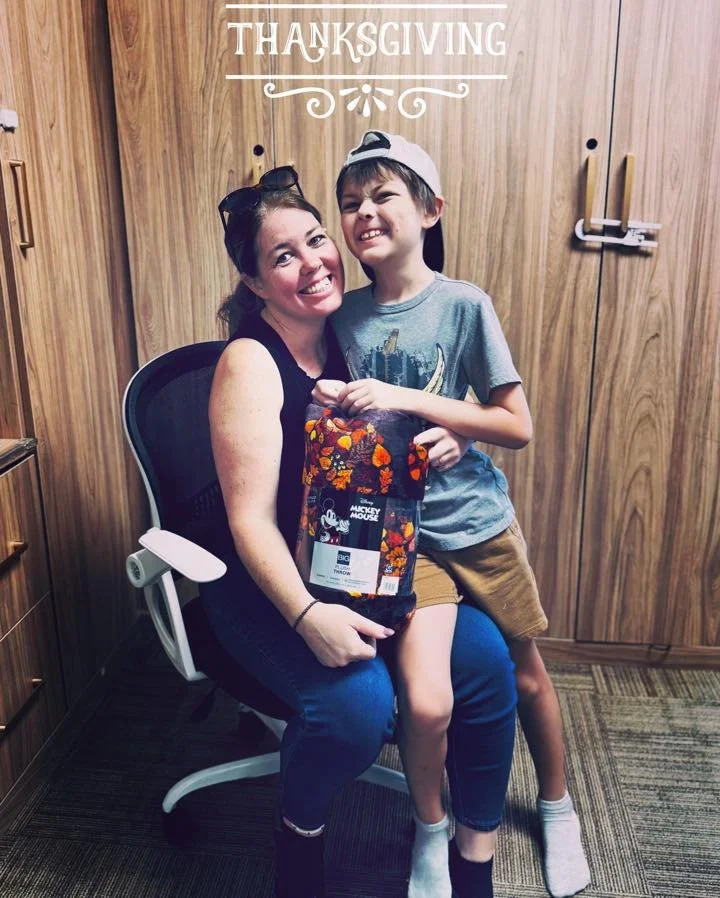 A woman and a young boy sitting on her lap, smiling, in front of a wooden wall with a 'Thanksgiving' sign, holding a gift bag, celebrating Thanksgiving.