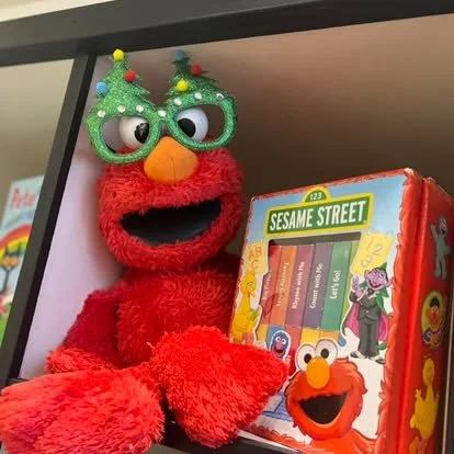 Elmo puppet wearing green glasses and Christmas tree-shaped sunglasses, sitting near a Sesame Street book.