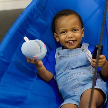 Smiling young boy sitting in a blue chair holding a plush toy and a remote control.