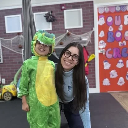 A young girl dressed in a green and yellow dinosaur costume standing next to a woman with long dark hair and glasses. They are inside a classroom decorated with Halloween-themed craft projects and decorations.