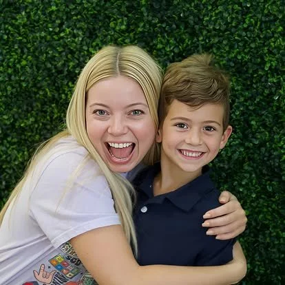 A woman and a young boy smiling and hugging in front of a green hedge wall.