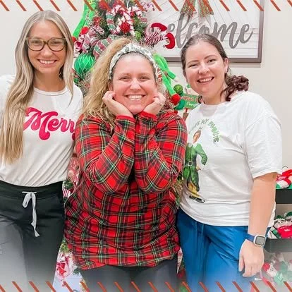 Three women smiling in front of Christmas decorations, including a decorated tree, holiday signs, and festive ornaments.