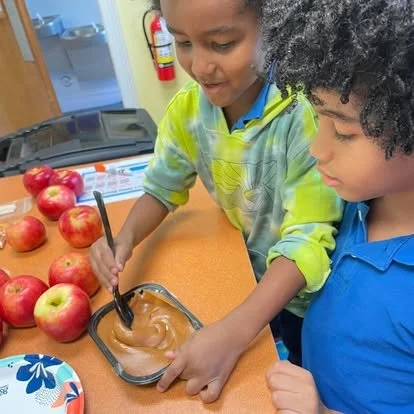 Two children stirring caramel sauce in a glass dish at a table with red apples.