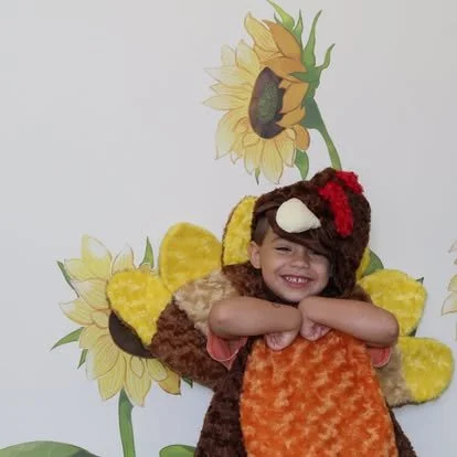 Child dressed in a sunflower-themed costume with a smiling face and arms crossed in front of a sunflower mural.