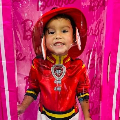 A young girl smiling inside a pink Barbie-themed play tent, wearing a red firefighter costume with a badge, and a helmet with a clear visor.