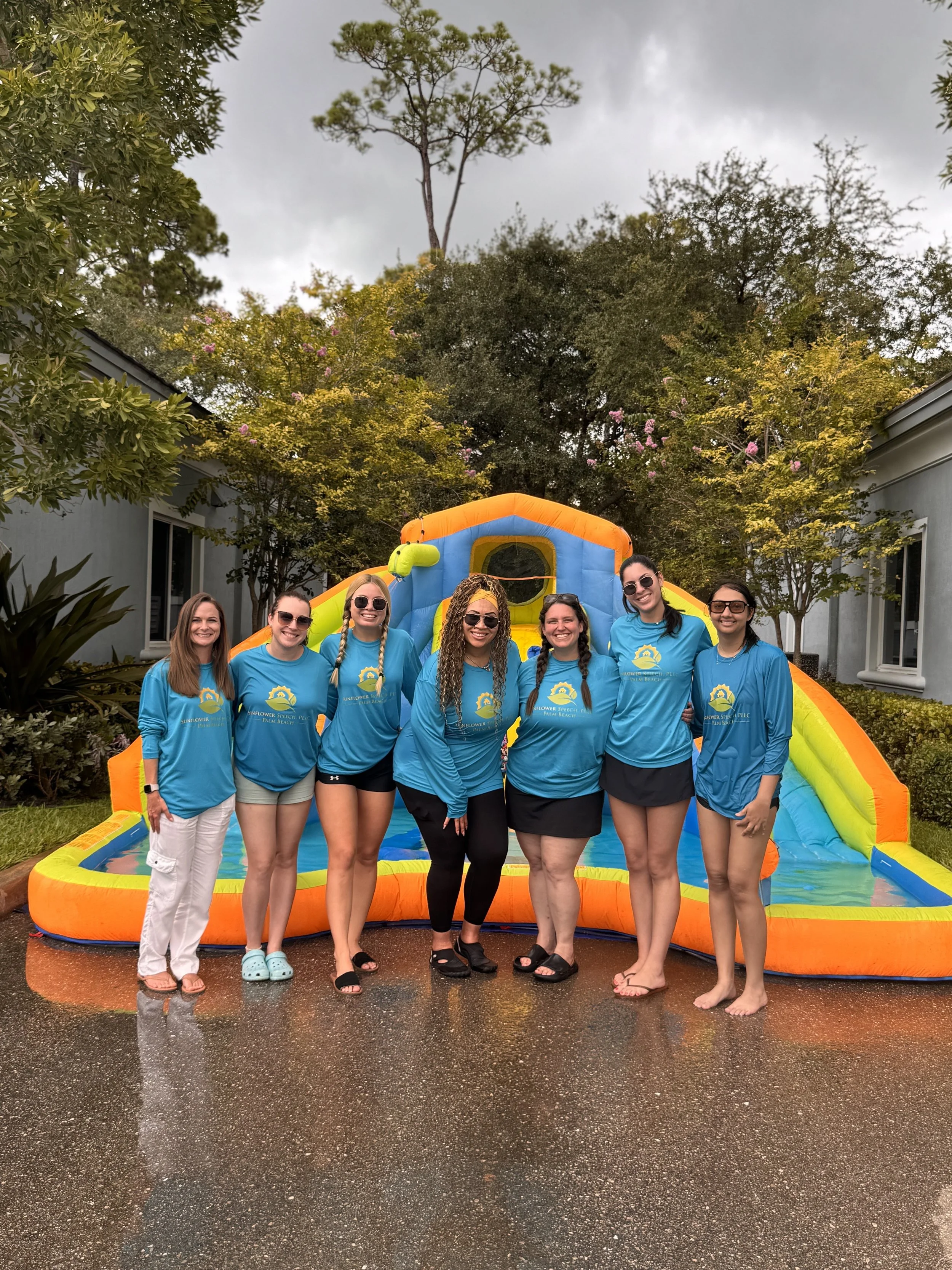 Seven women standing in front of an inflatable water slide, smiling, at a beach or outdoor event, all wearing matching blue shirts with a logo or design.