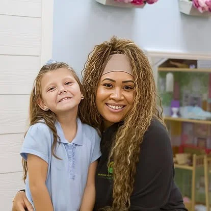 A woman with curly hair and a young girl in a light blue shirt smiling and hugging each other indoors.