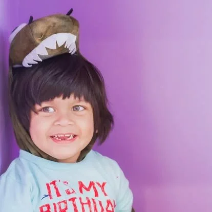 Child wearing a T-Rex costume hat with a purple background.