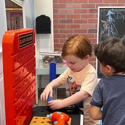 Two young boys playing with a red Connect Four game, one placing a disc into the game while the other watches, in a classroom setting with a chalkboard and bookshelf in the background.