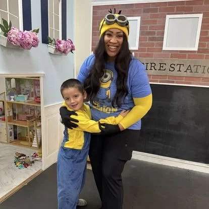 A woman and a young boy dressed in Minion costumes, smiling and hugging each other in an indoor setting with pink flowers and a miniature play shop in the background.