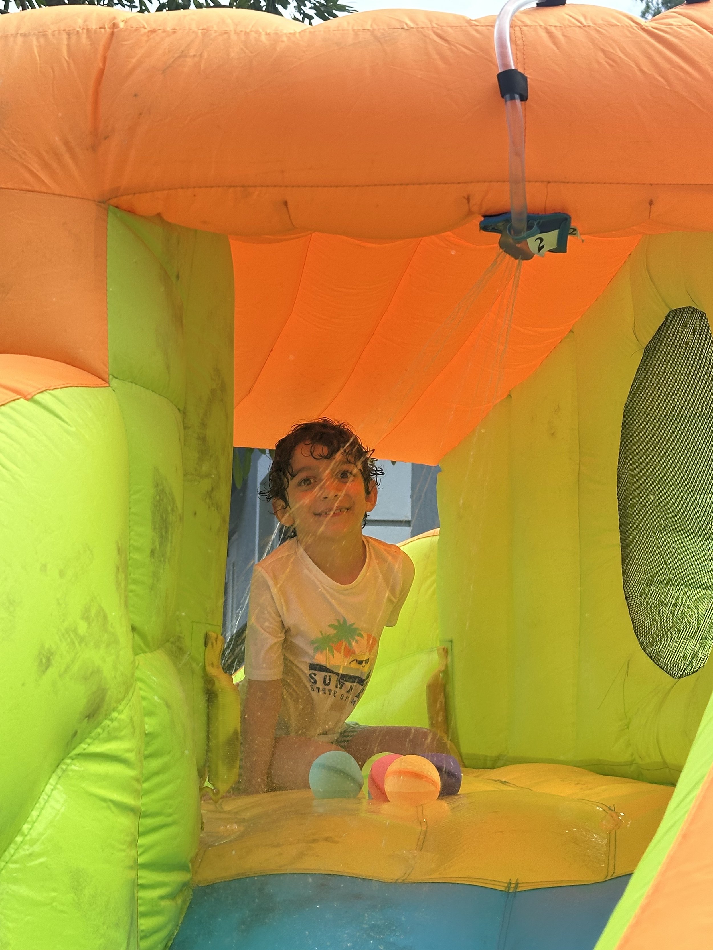 A young boy with curly hair inside a colorful inflatable water bounce house, smiling and playing with pink, purple, blue, and orange balls.
