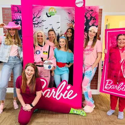 Group of seven women dressed as Barbie characters, posing at a Barbie-themed event with pink decorations, a large pink frame, and Barbie-themed posters.
