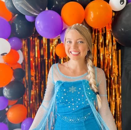 Woman dressed as Elsa from Frozen in front of Halloween-themed decorations with black, purple, orange balloons, and a shiny orange and black curtain.