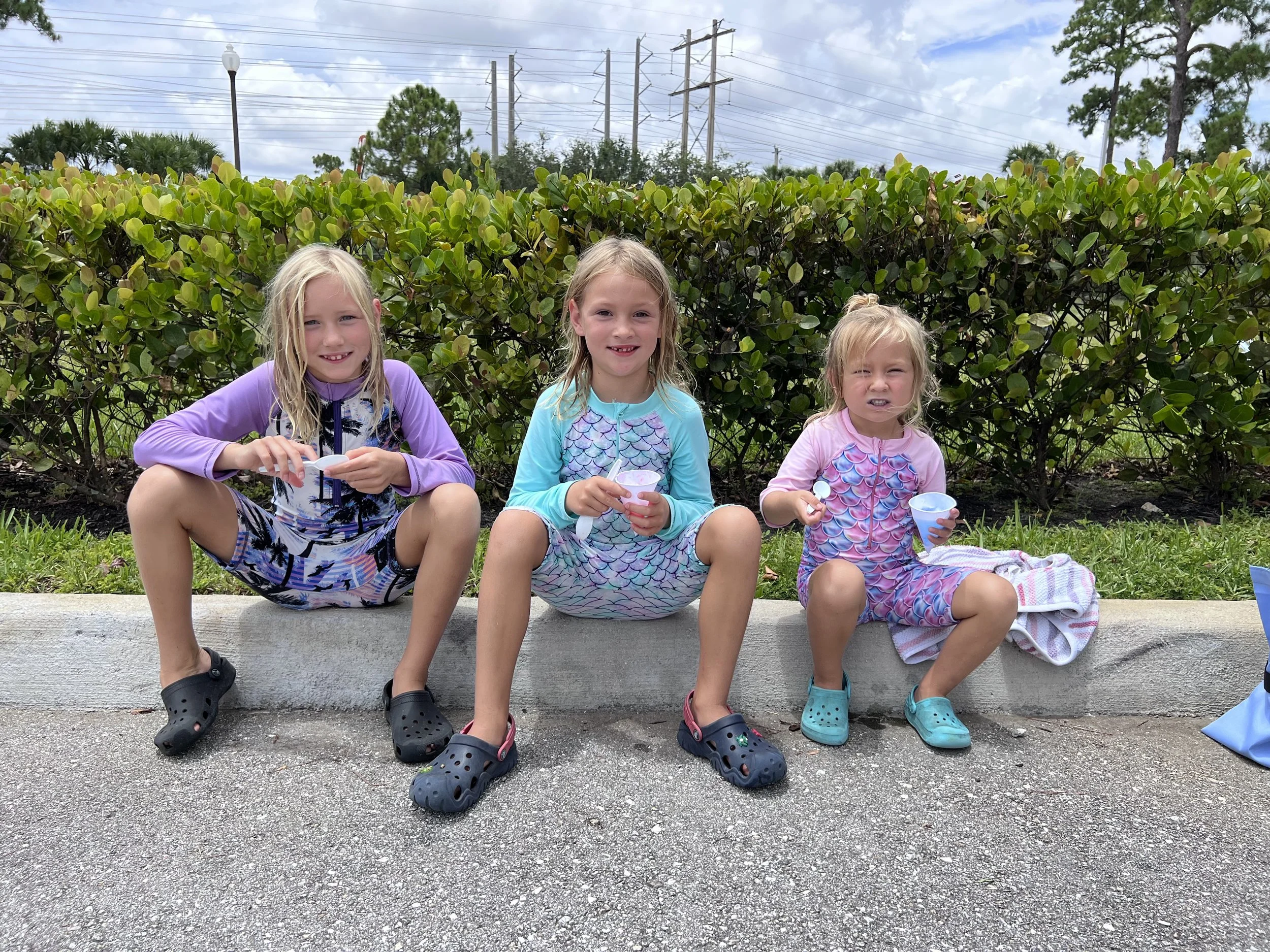 Three young girls sitting on a curb, holding cups and spoons, wearing colorful swimsuits, with bushes and trees in the background on a cloudy day.