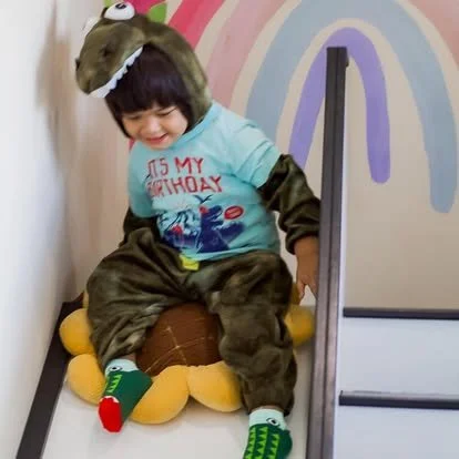 Child in a dinosaur costume sitting on a yellow toy dinosaur on a slide at a birthday party.